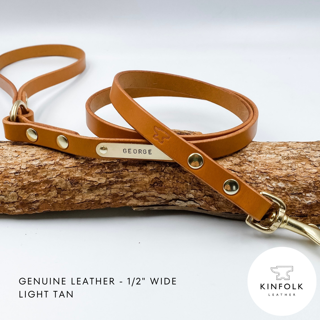 Light tan leather dog leash with gold hardware and nameplate on a wooden branch, branded 'KINFOLK LEATHER'.