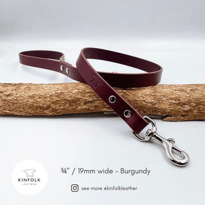 A burgundy leather dog leash with silver hardware, draped over a piece of natural wood on a light gray background. Text at the bottom reads '3/4" / 19mm wide - Burgundy' with the Kinfolk Leather logo and social handle.