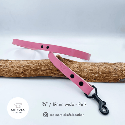 A pink leather dog leash with black hardware and rivets, laid diagonally across a natural wood branch on a white studio background. Text at the bottom reads '3 3/4" / 19mm wide - Pink' with the Kinfolk Leather logo and social handle.