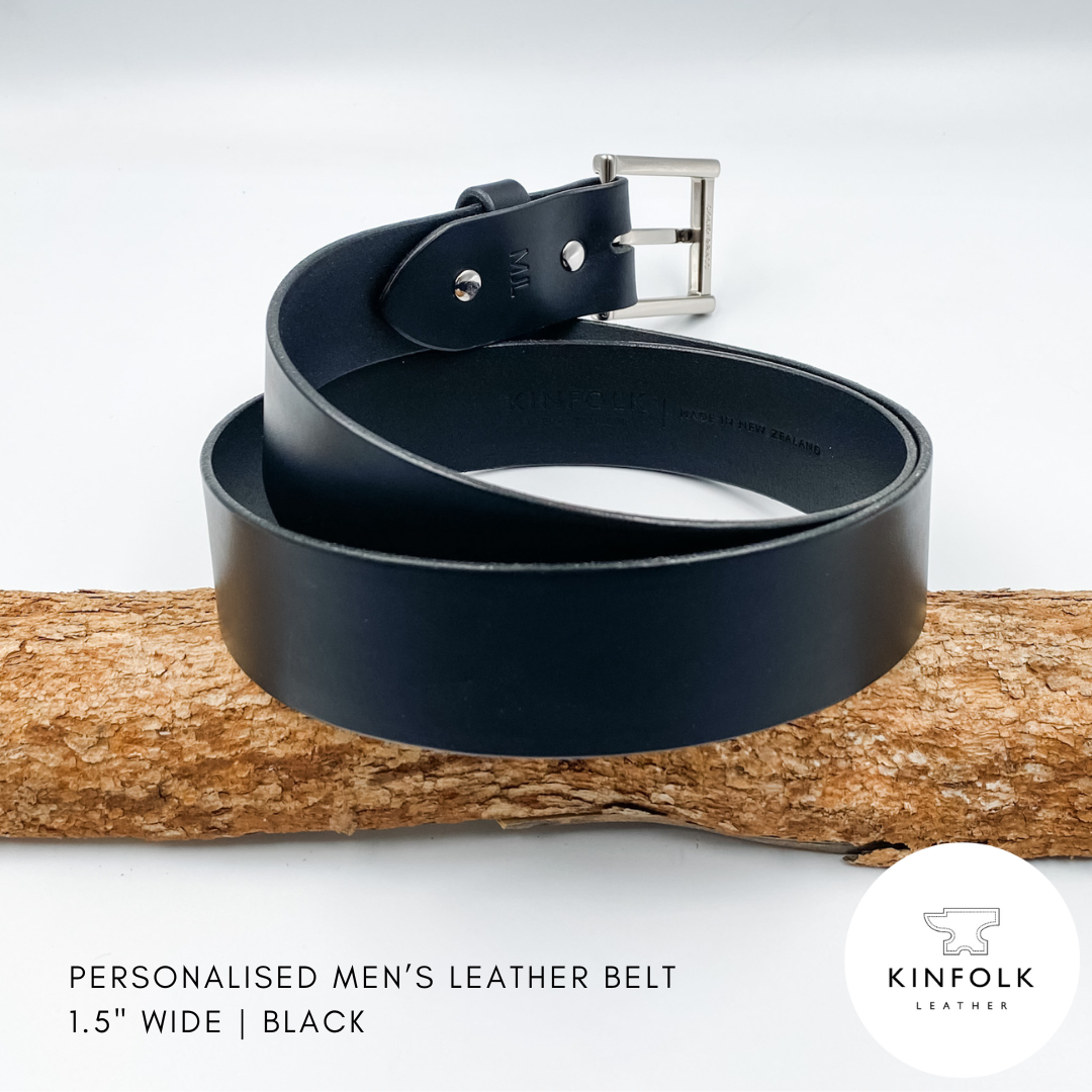 Black leather belt with silver buckle on a wooden log against a white background, featuring the Kinfolk Leather brand.